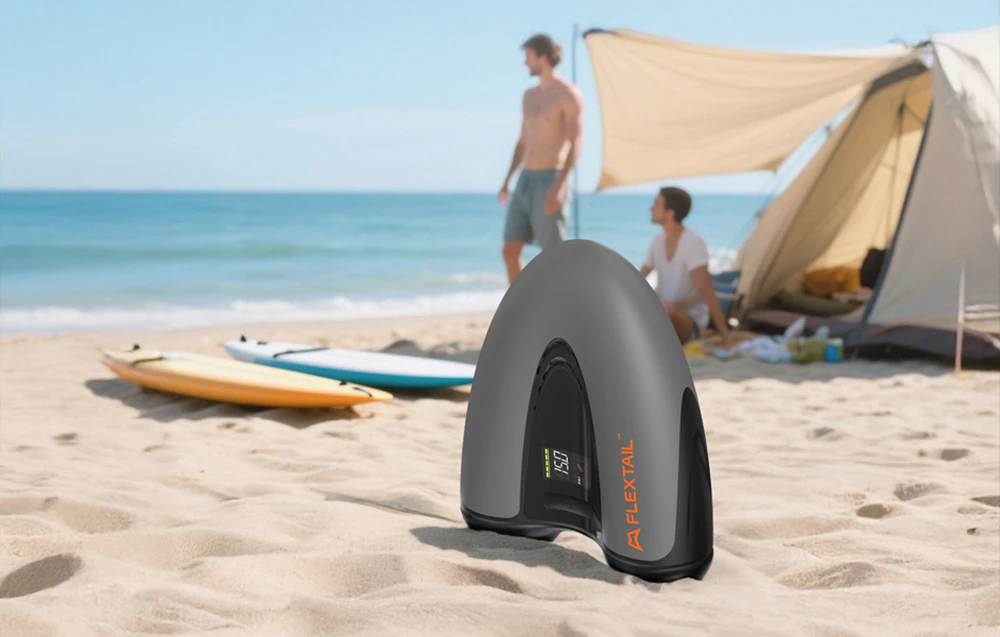 Pompa portabilă Flextail Evo SUP Pump Pro se află pe plajă lângă un cort și plăci SUP.