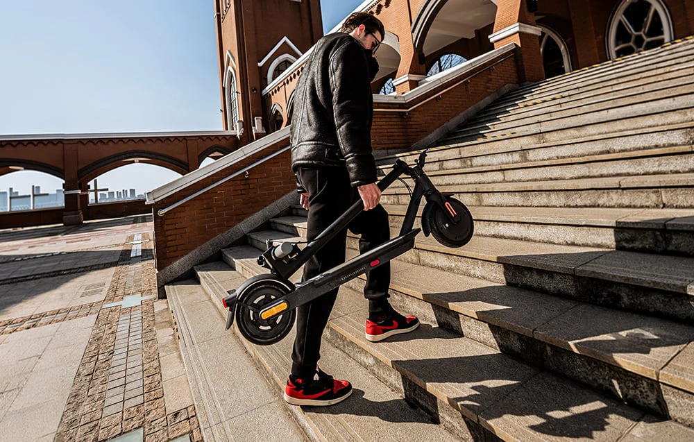 Un bărbat cară un scuter electric pliat pe treptele largi de piatră din fața unei clădiri din cărămidă.