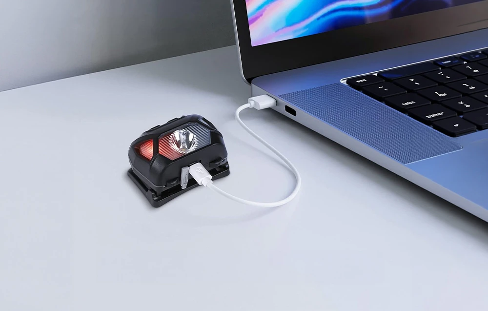 O lanternă mică, neagră, cu un LED roșu, se află pe un birou, conectată la un laptop printr-un cablu USB alb.