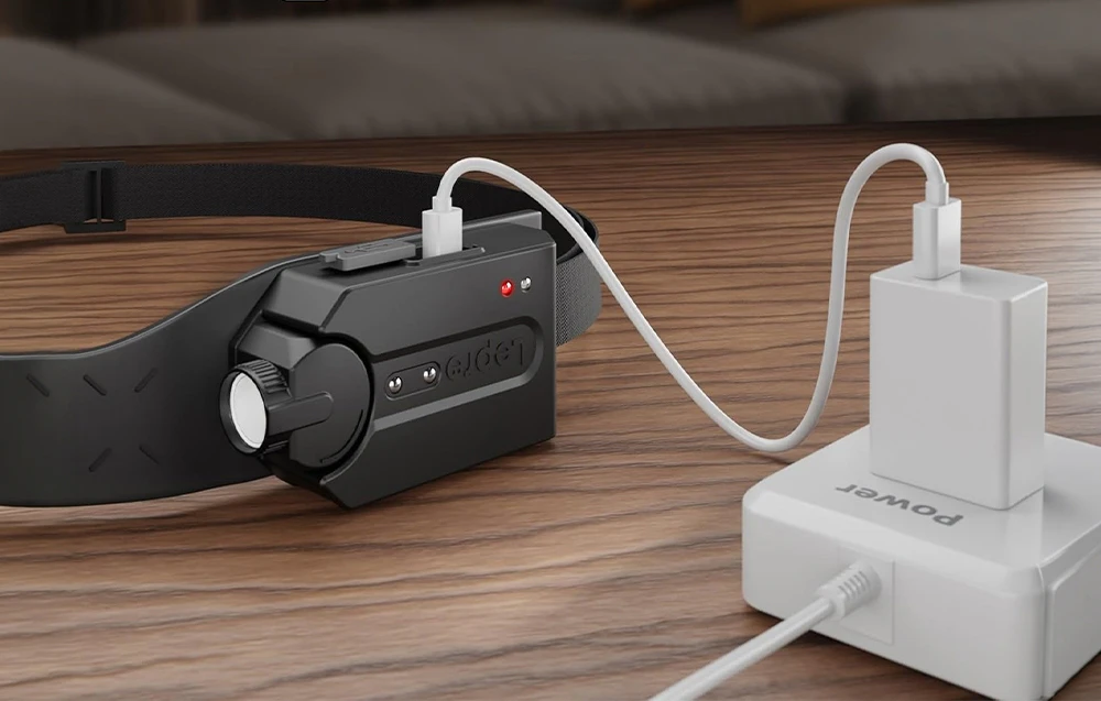 O lanternă frontală neagră conectată printr-un cablu USB alb la un încărcător de perete așezat pe un blat de lemn.