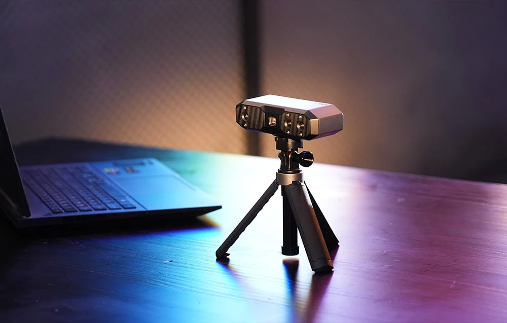 Dispozitivul Revopoint Mini 2 pe un trepied lângă un laptop, iluminat de lumină colorată într-un birou modern