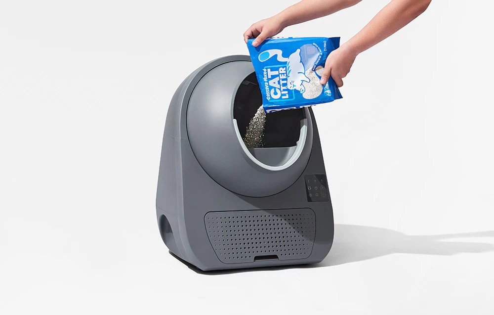 O persoană care toarnă nisip pentru pisici dintr-un pachet albastru „Cat Litter” într-o cutie automată de nisip de culoare gri