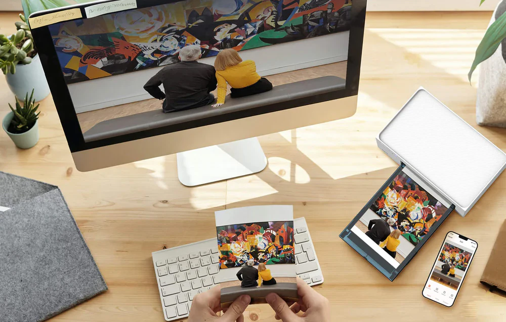 Un birou cu un monitor, un smartphone și o imprimantă Liene; mâini care țin o fotografie imprimată dintr-o galerie de artă.