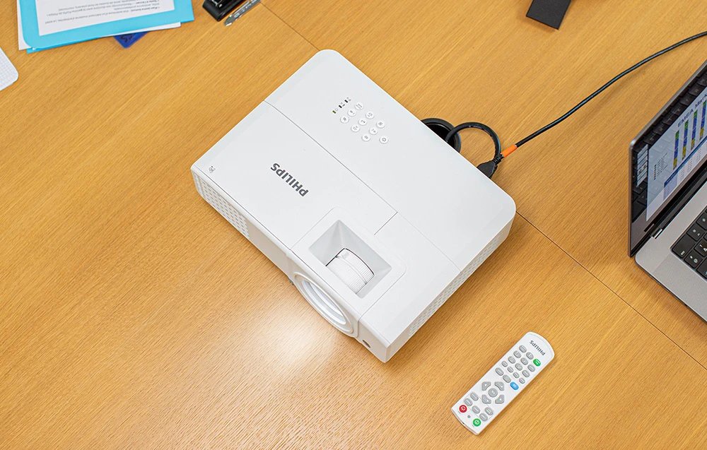 Vedere de sus a unui proiector PHILIPS așezat pe o masă lângă o telecomandă, un laptop și documente