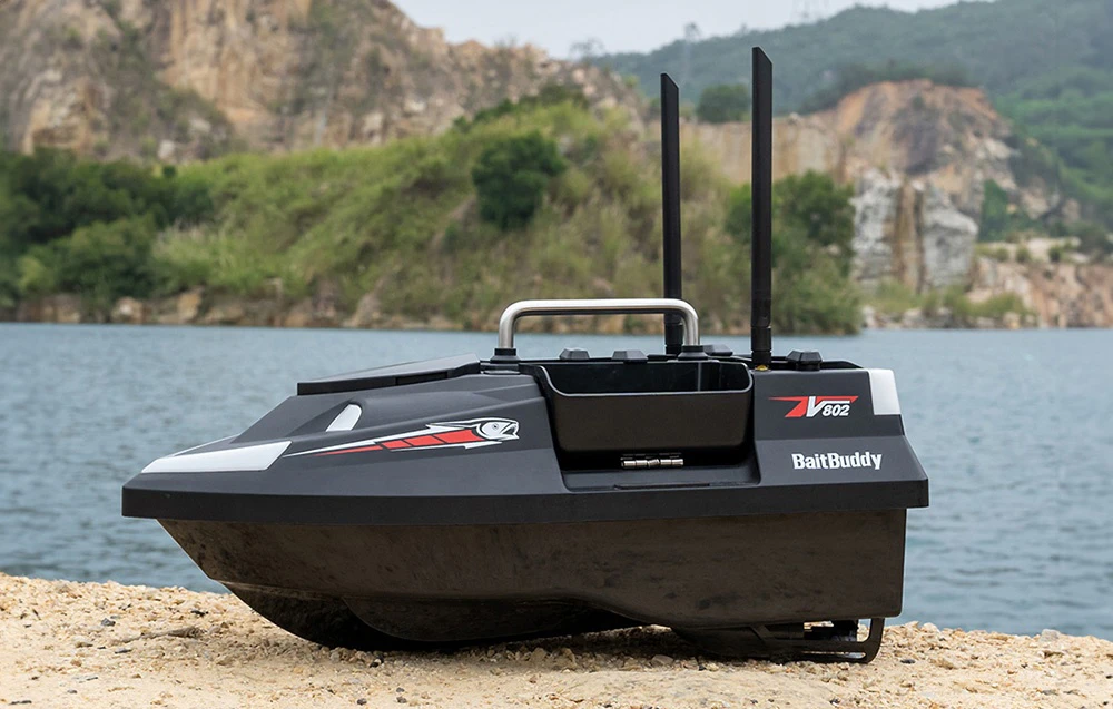 Barca de momeli RC BaitBuddy V602 cu două antene care stă pe malul lacului cu munții în fundal.