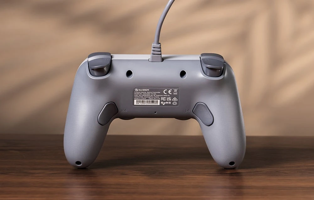 Partea din spate a controlerului GameSir gri, cu butoanele, eticheta și cablul vizibile pe masa de lemn.
