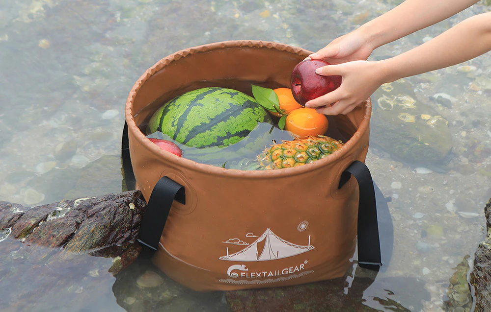 O găleată pliabilă de camping scufundată într-un lac, folosită pentru răcirea fructelor: pepene verde, ananas și mere
