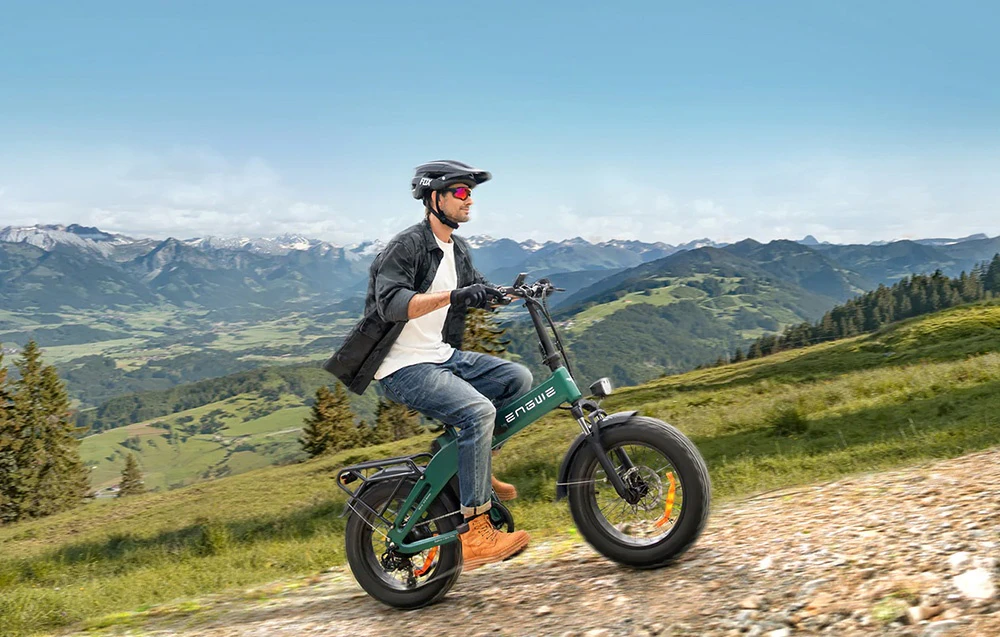 Un bărbat care poartă cască conduce o bicicletă electrică pliabilă verde pe un traseu montan, având în fundal o panoramă montană
