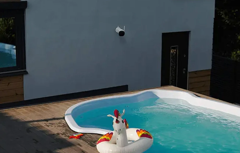 O piscină în curtea din spate cu un unicorn gonflabil, cu un perete alb al clădirii în fundal, pe care este montată o cameră de securitate neagră