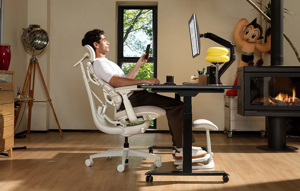 O persoană stă așezată pe un scaun ergonomic alb lângă un birou cu ecran și ține un smartphone în mână lângă șemineu