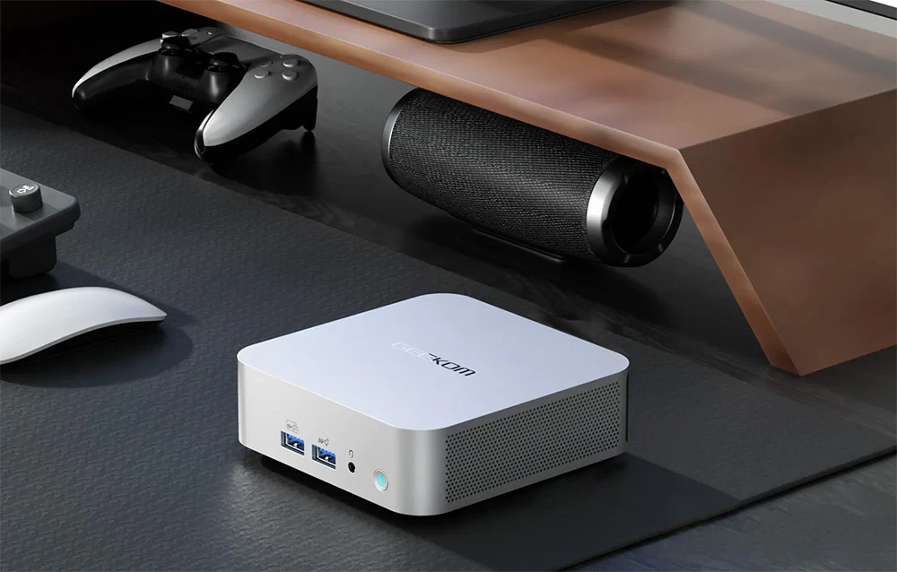 Un mini PC argintiu pe o suprafață de birou întunecată, lângă un difuzor, un mouse și un controler de jocuri.