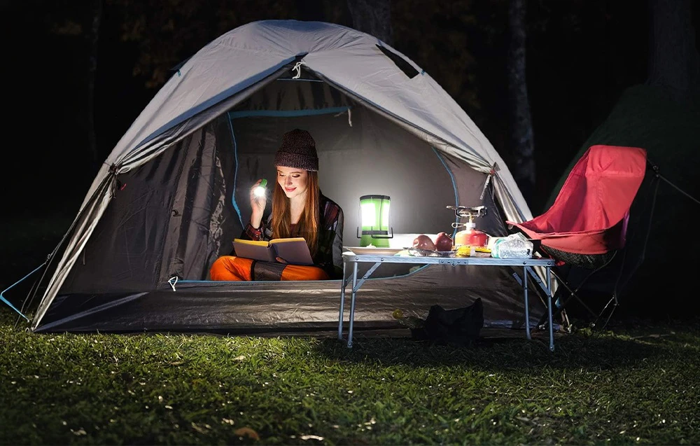 O femeie citește o carte în interiorul unui cort gri; o lampă de camping verde stă pe o masă pliantă lângă ea.