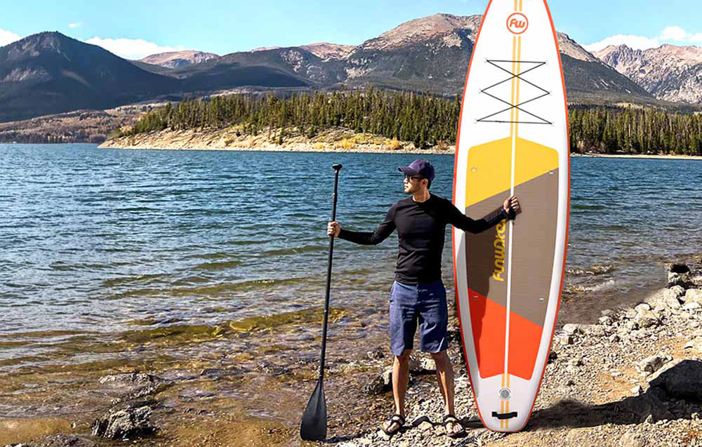Bărbat cu șapcă de baseball stând pe malul unui lac, ținând o vâslă și o placă de surf, cu munții în fundal
