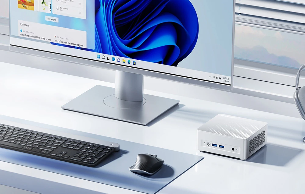 Mini PC argintiu pe un birou alb, lângă un monitor Windows, un mouse negru și o tastatură