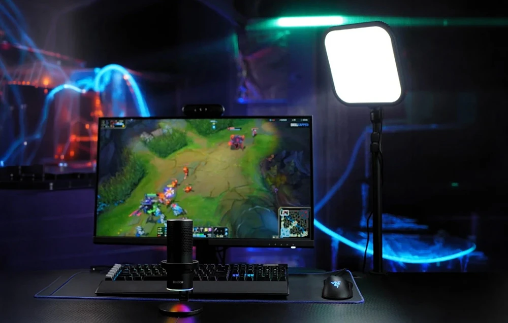 Un microfon Godox, o tastatură, un mouse Razer, un monitor aprins și o lampă strălucitoare pe un birou într-o cameră întunecată cu iluminare colorată