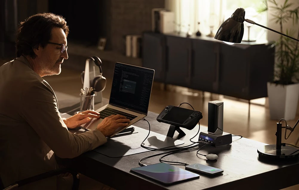Un bărbat care poartă ochelari lucrează la un laptop. Lângă el, pe birou, o consolă, o tabletă, un smartphone și căști wireless sunt conectate la un powerbank cu stație.