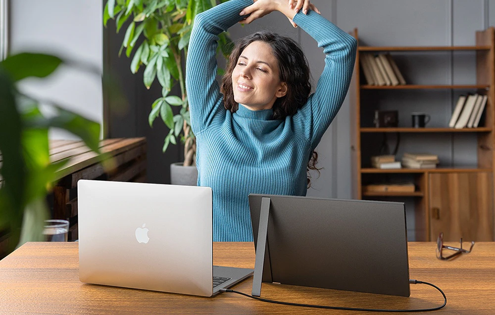 O femeie zâmbitoare într-un pulover stă la un birou pe care se află un laptop și un monitor portabil conectat la acesta.