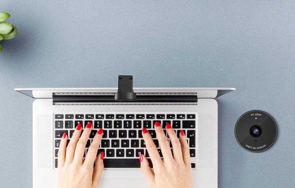 O vedere de sus în jos a mâinilor care tastează pe tastatura unui laptop, pe care este montată o cameră mică, neagră.