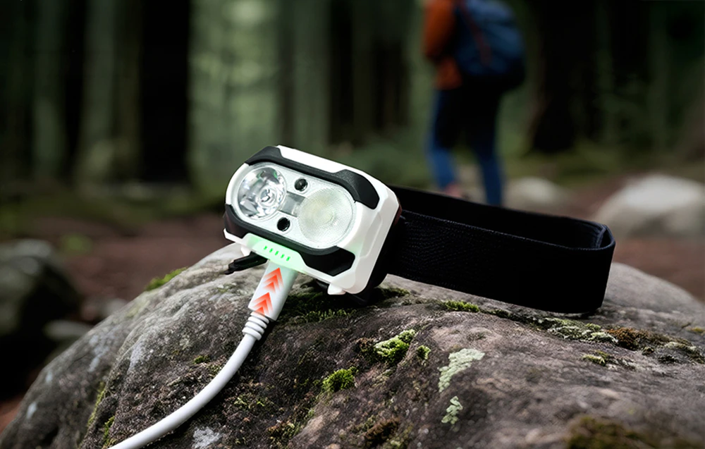 O lanternă frontală încărcată printr-un cablu USB alb; silueta neclară a unui turist în pădure în fundal.