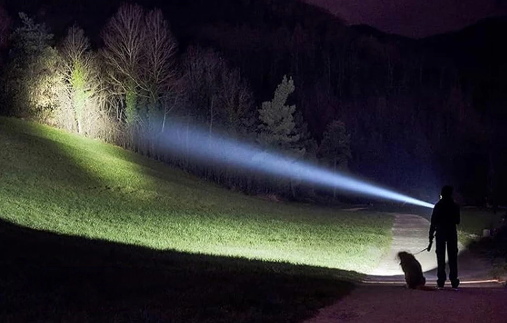 O persoană care plimbă un câine pe o potecă iluminată de fasciculul alb puternic al lămpii pe versantul unui munte.