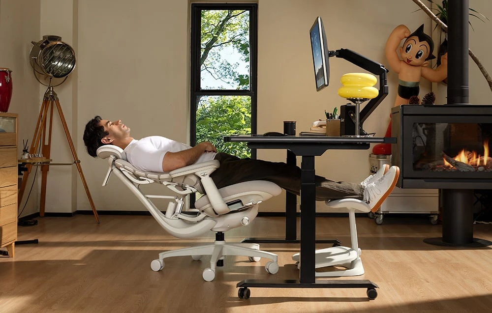 O persoană este întinsă pe un scaun ergonomic alb lângă un birou cu un ecran și un șemineu