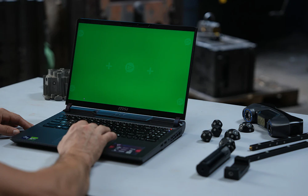 O persoană operează un laptop cu un ecran verde, componentele sistemului de scanare 3D aflându-se lângă acesta