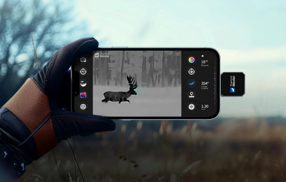 Un smartphone conectat la o mică cameră cu imagistică termică afișează o imagine alb-negru a unui cerb într-o poiană.