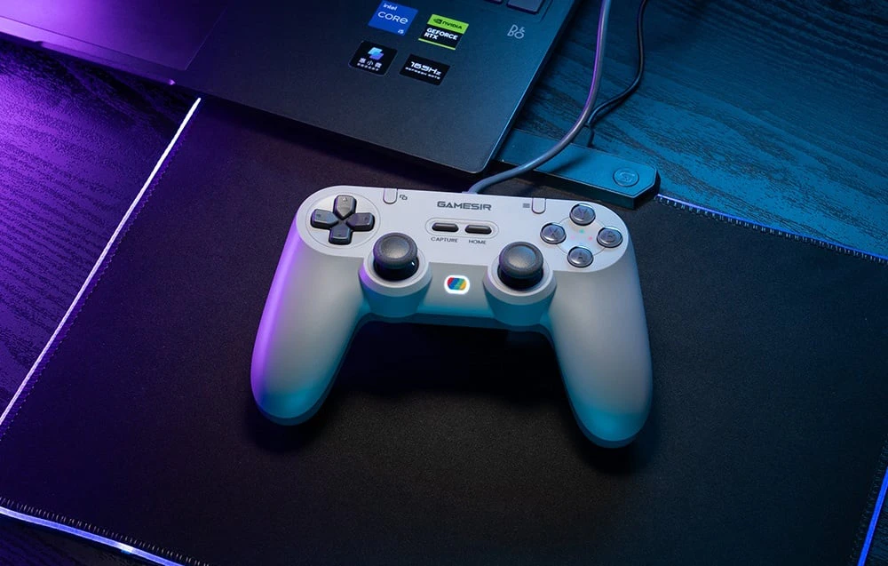 Un controler GameSir gri așezat pe un suport în fața unui laptop, iluminat de o lumină albastră-violetă.