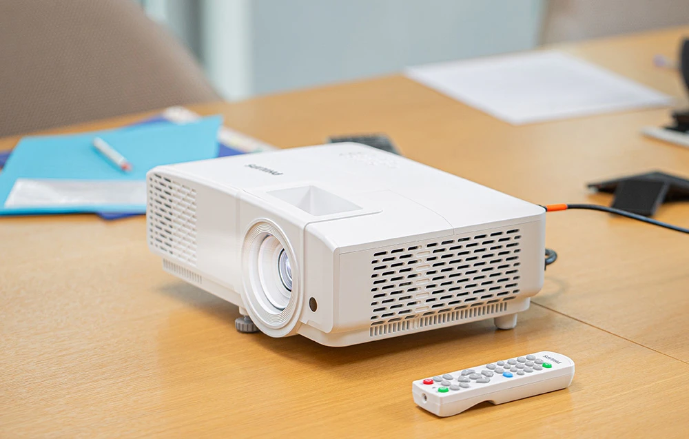 Un proiector PHILIPS alb se află pe o masă de conferințe din lemn, lângă o telecomandă albă