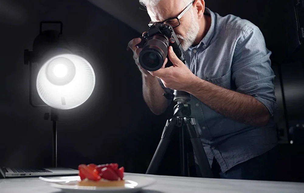 Un fotograf care face o fotografie unui desert pe o farfurie, iluminat de lumina albă și strălucitoare a lămpii.