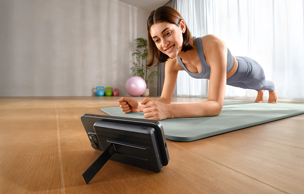 Femeia face exerciții pe un covor în cameră. În fața ei se află un smartphone conectat la un powerbank magnetic cu un suport.