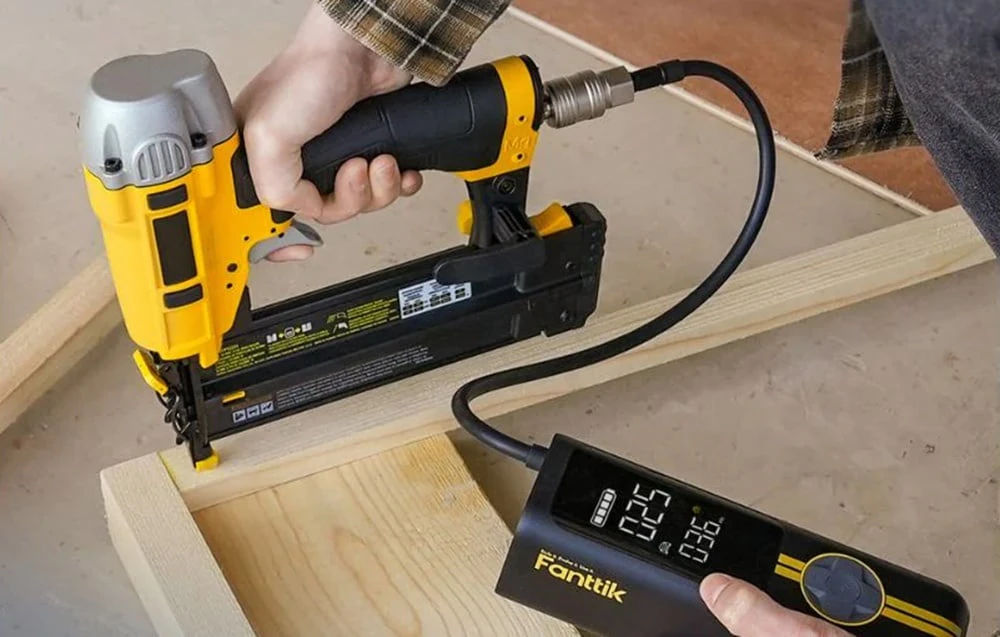 O persoană folosește un cuier pneumatic galben și negru conectat la un compresor portabil cu afișaj digital, atașând piese din lemn.