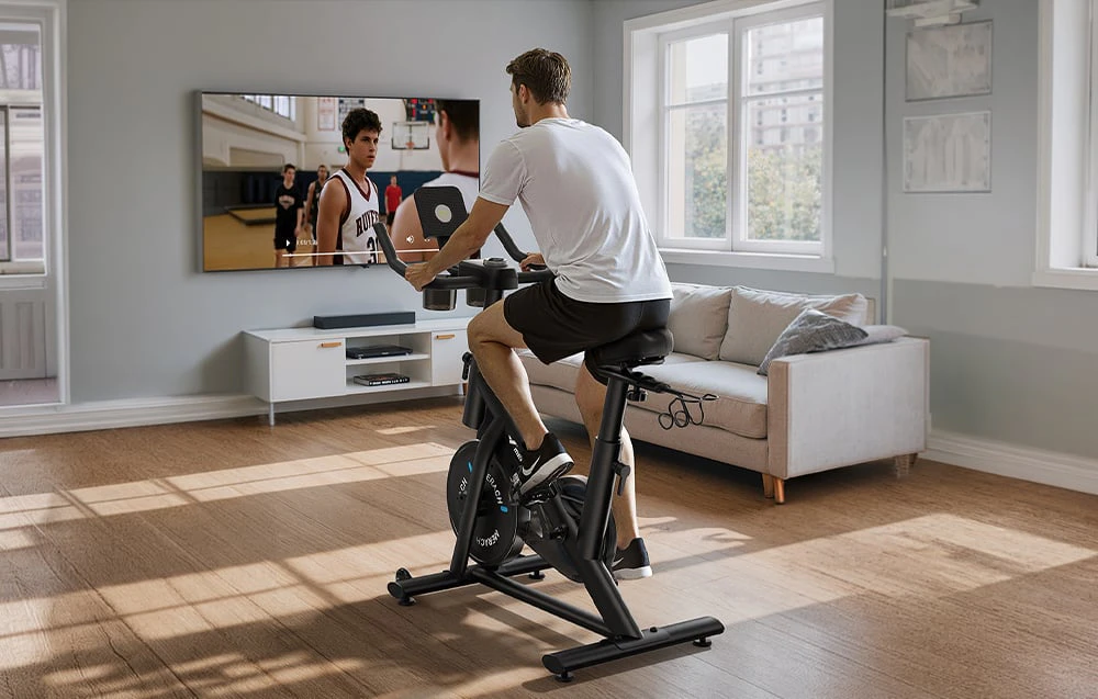 Bărbat care face exerciții fizice pe o bicicletă staționară și se uită la un meci la televizor
