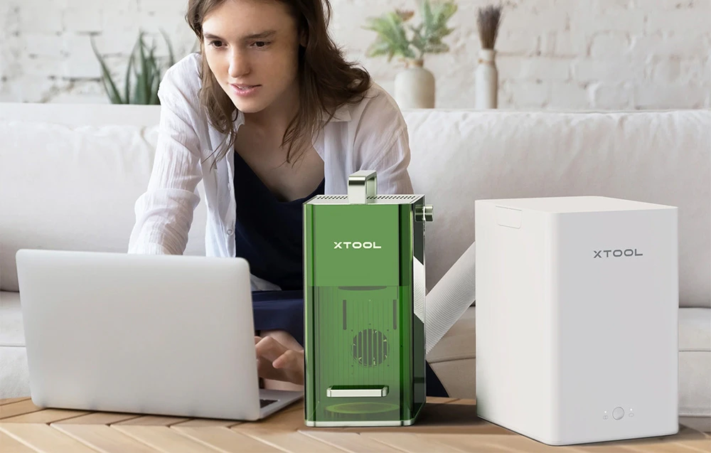 O femeie care lucrează la un laptop lângă un laser verde și un aspirator de fum xTool alb așezate pe o masă.