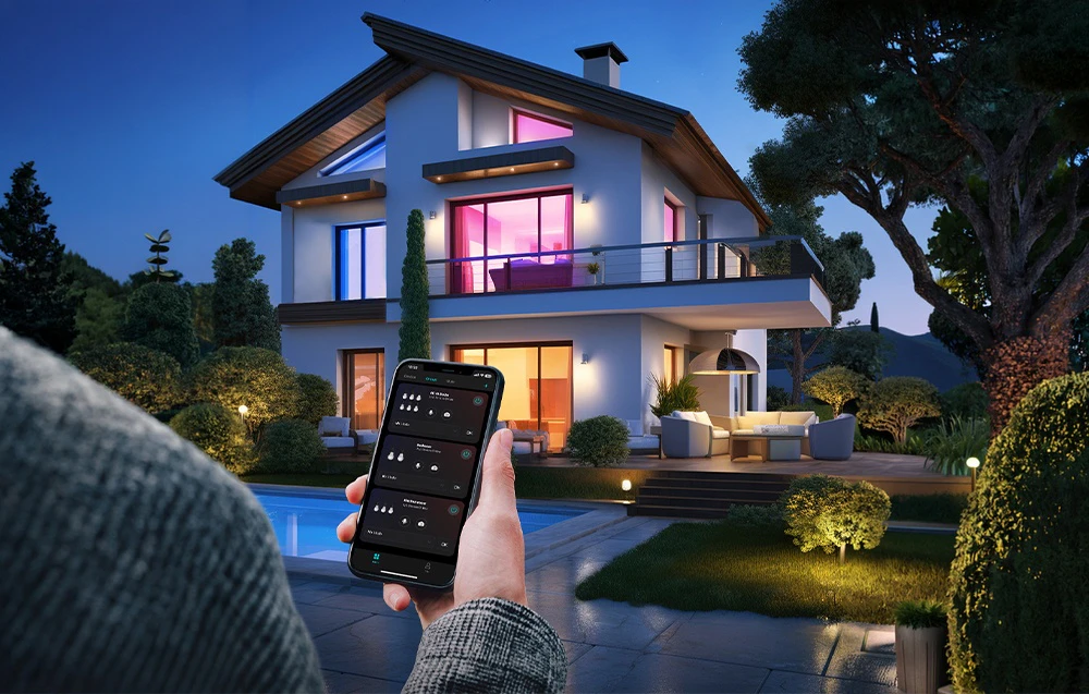 O mână ține un smartphone cu o aplicație de control al iluminatului deschisă, pe fundalul unei case unifamiliale moderne la apus.