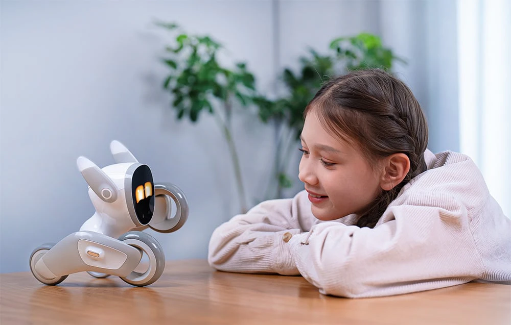 O fetiță zâmbitoare, cu codițe și un hanorac roz, stă rezemată de o masă și se uită la robotul interactiv Loona