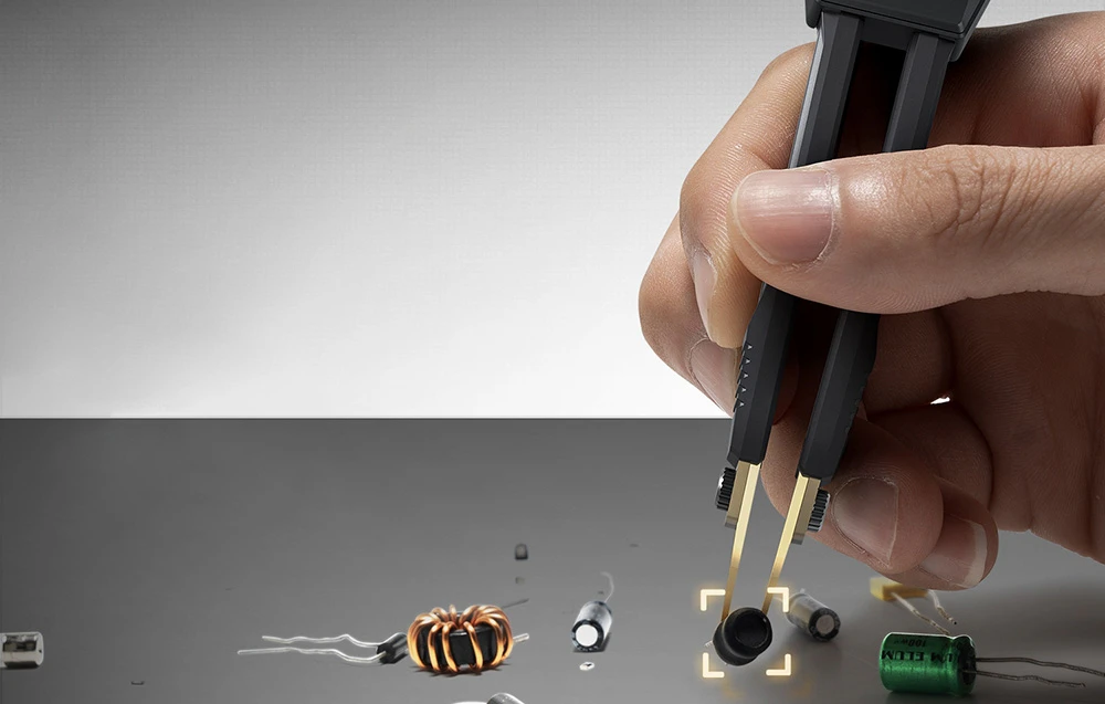 O mână ține o pensetă electronică, care prinde o componentă electronică mică neagră dintre componentele împrăștiate.