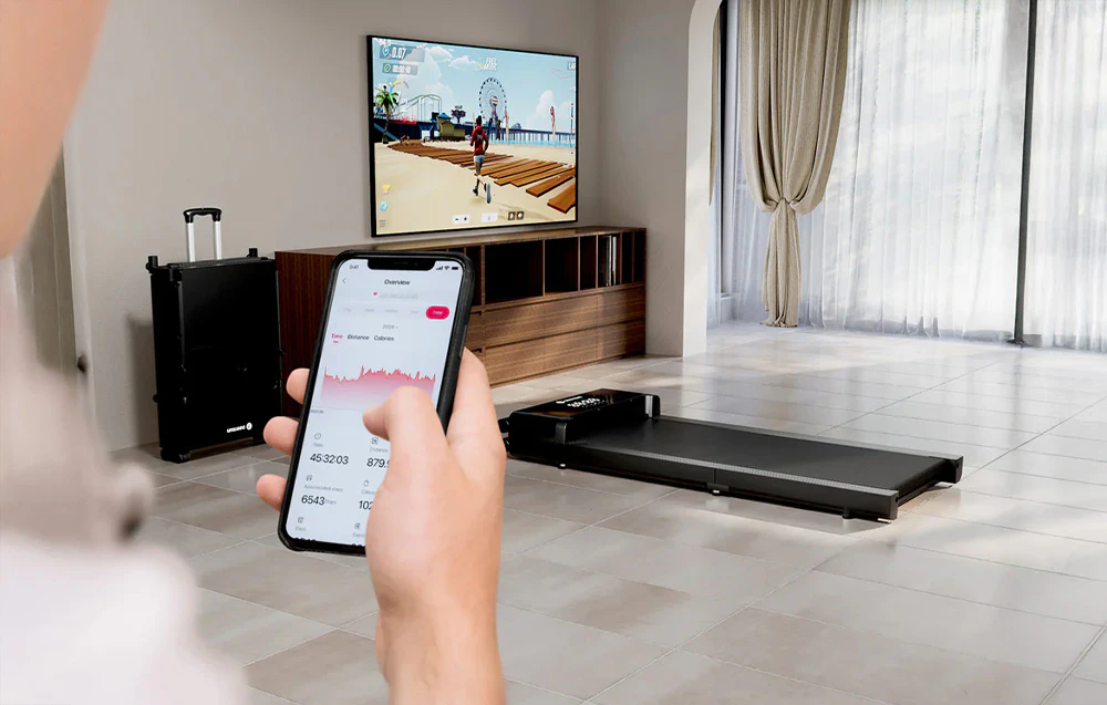 Mâini ținând un smartphone cu o aplicație de fitness pe fundalul unei camere de zi cu o bandă de alergare și un televizor