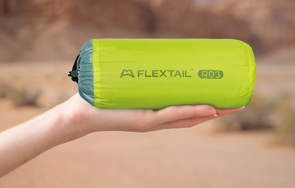 O mână care ține covorașul de camping Flextail R03, rulată și ultracompactă, pe fundalul unui peisaj estompat