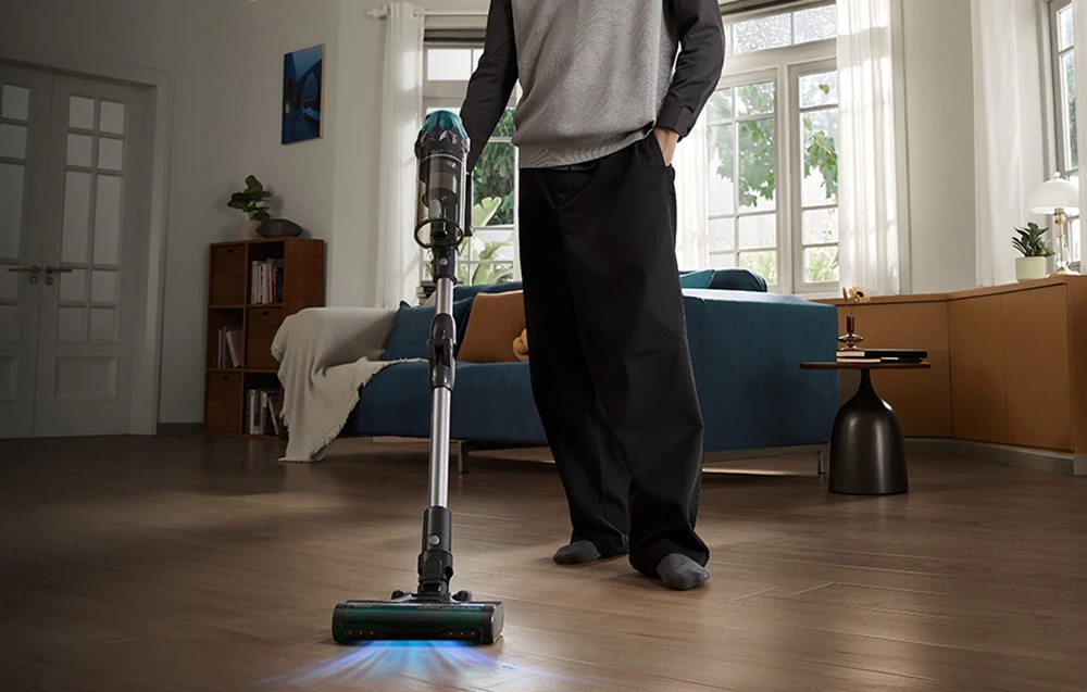 Un bărbat stând în sufragerie cu un aspirator vertical fără fir, cu iluminare de fundal albastră aprinsă