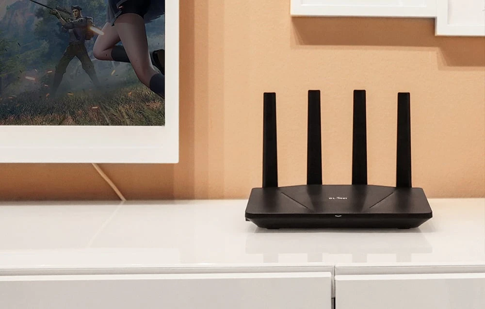 Un router negru așezat pe un blat alb lângă o porțiune a unui televizor de culoare deschisă, pe fundalul unui perete bej dintr-o cameră