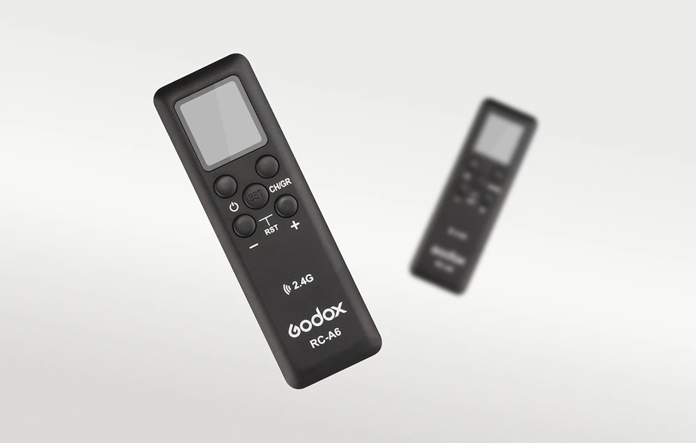 Telecomanda neagră Godox RC-A6 cu ecran pe un fundal deschis, cu o a doua telecomandă estompată în fundal