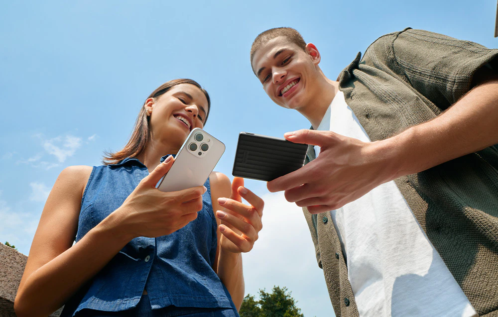 O femeie zâmbitoare ține în mână un smartphone alb. Lângă ea este un bărbat zâmbitor care ține în mână un powerbank negru.