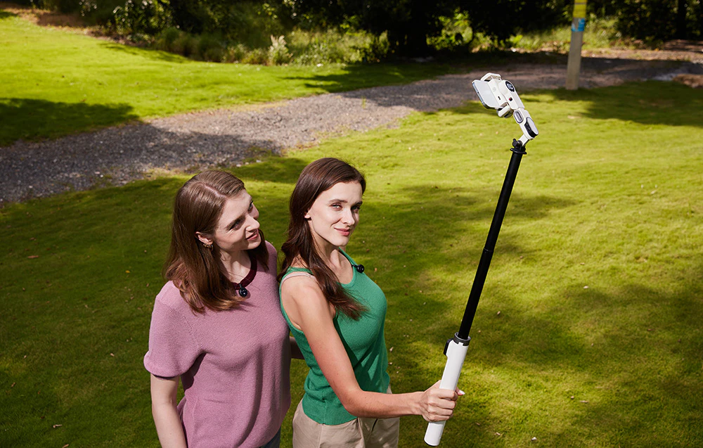 Două femei stau pe gazon și își fac un selfie cu un gimbal ținut deasupra capetelor.