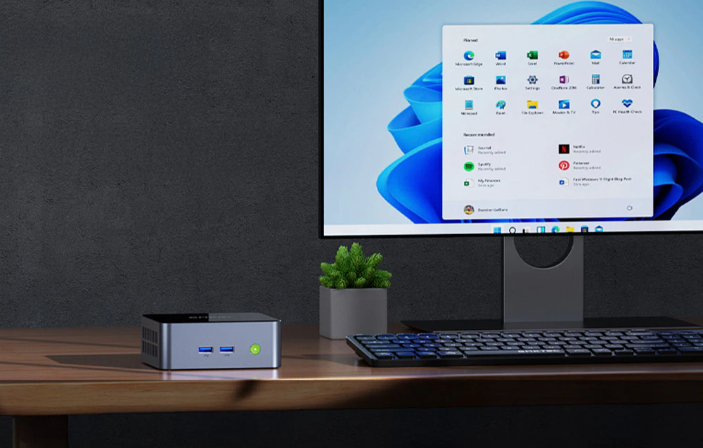 Mini PC GMKtec pe birou, lângă monitorul Windows 11, tastatură și ghiveci de flori