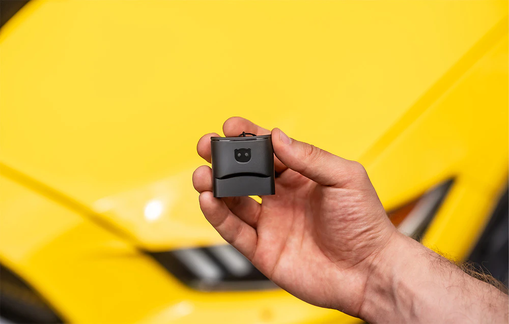O mână prezentând adaptorul compact Carly OBD2 pe fundalul unei mașini sport galbene