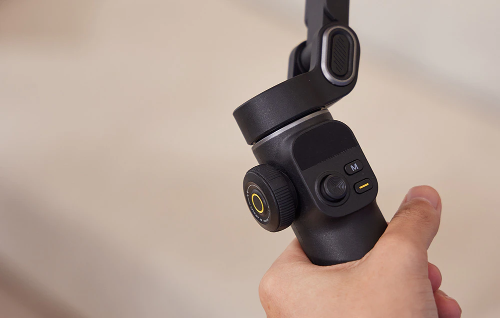 O mână care ține un gimbal negru — prim-plan al panoului de control cu un cadran și butoane.