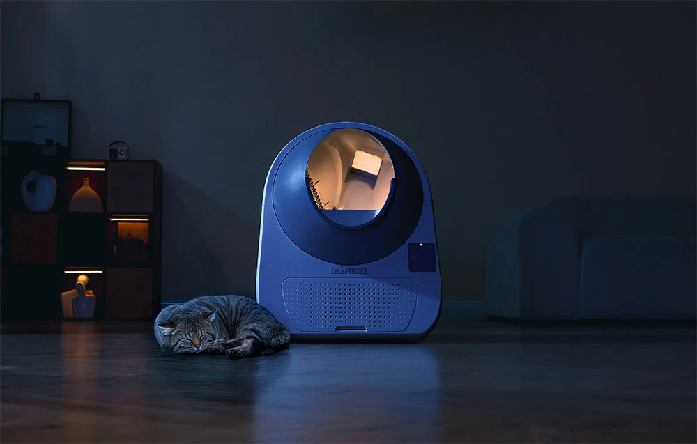 O pisică care doarme pe podea lângă o cutie de nisip automată CatLink iluminată din spate, într-o cameră întunecată