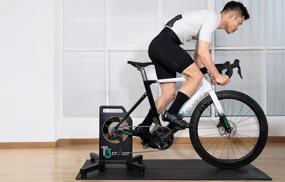 Un bărbat se antrenează pe o bicicletă albă montată pe un trainer Cycplus T3 într-un interior luminos