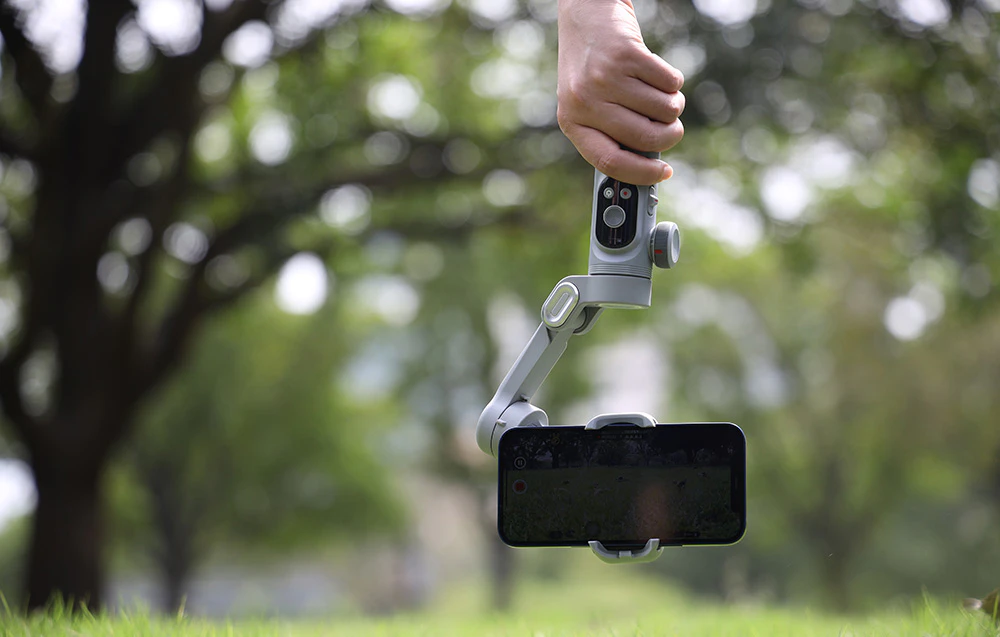 O mână ține un gimbal cu un smartphone îndreptat în jos în timp ce înregistrează în aer liber.